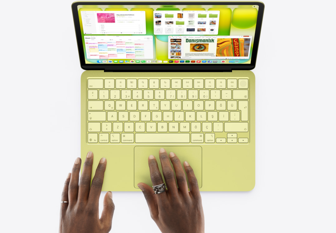 MacBook Neo, puslu sarı renk, bir kişinin eli trackpad’in üzerinde duruyor, Müzik, Takvim ve Microsoft PowerPoint uygulamaları gibi birçok uygulama penceresi açık
