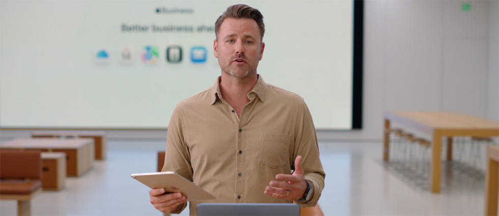 Presenter speaks while referencing an iPad, and a large presentation screen displays various business app icons.